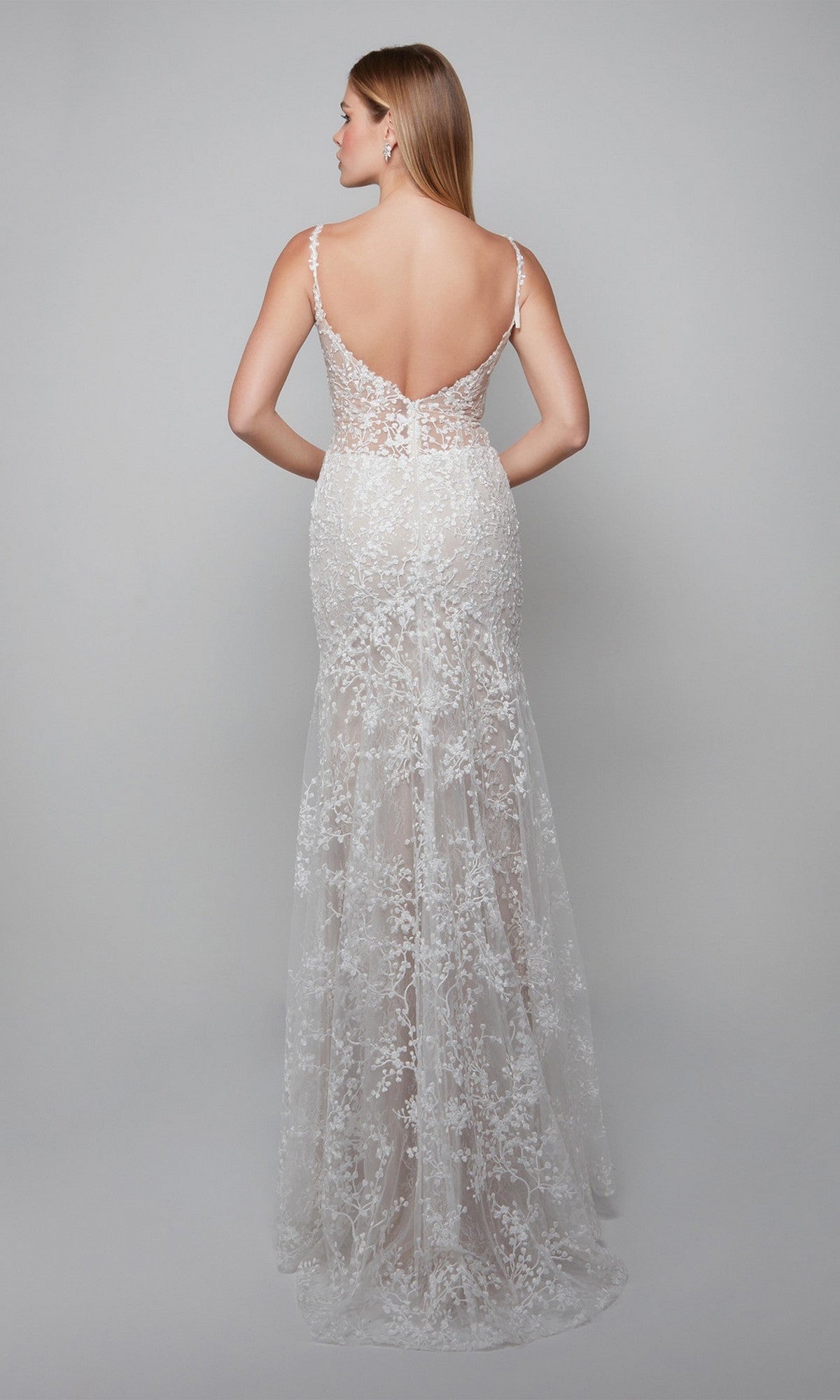 A woman stands with her back to the camera in the Alyce 7077 long white dress, featuring a sleeveless design, lace appliques, an ivory hue, and a deep V-back against a plain background.